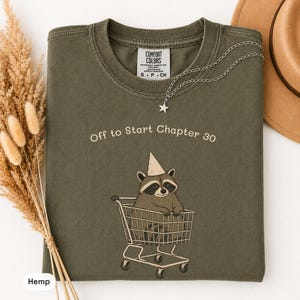 May include: An olive green crew neck t-shirt featuring a graphic of a raccoon wearing a party hat while sitting in a shopping cart. The text above the raccoon reads, Off to Start Chapter 30. The shirt has a Comfort Colors brand label inside.