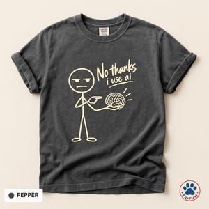 Peut inclure: Un t-shirt à manches courtes gris anthracite avec un motif crème représentant un bonhomme allumette pointant un cerveau. Le texte "No thanks i use ai" est imprimé au-dessus du cerveau. Le t-shirt a un col rond et une étiquette Comfort Colors.