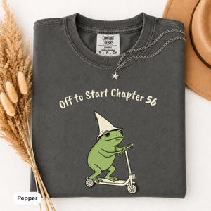 May include: A charcoal gray t-shirt featuring a graphic of a green frog wearing a pointed hat and riding a scooter. The text above the frog reads "Off to Start Chapter 56" in a light cream color. The shirt has a Comfort Colors brand label.