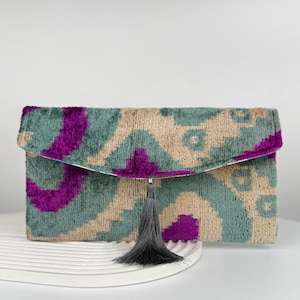 May include: A rectangular clutch bag featuring a velvet-like texture with an abstract pattern in shades of teal, deep purple, and beige. The envelope-style flap is centred with a dark grey decorative tassel hanging from the bottom edge.