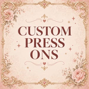 May include: A square sign with a light pink background featuring the dark red text CUSTOM PRESS ONS in the center. The sign is framed by an ornate gold border with floral accents in the corners and small heart and star icons throughout.