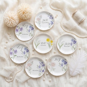 Op de afbeelding: Kleine witte decoratieve borden met bloemmotieven en gouden randen. Elk bord heeft initialen en de datum 08.18.2026. De bloemen zijn onder andere viooltjes, narcissen en rozen. Perfect voor huwelijksbedankjes of speciale evenementen.