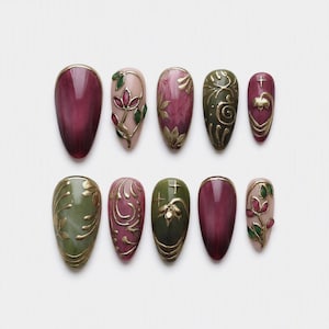Op de afbeelding: Een set van tien kunstnagels in amandel- en ovale vormen. De ontwerpen bevatten diep bordeauxrode, olijfgroene en zachtroze basissen met verhoogde gouden metalen filigraan, bloemmotieven en kleine steraccenten op de verschillende nageltips.