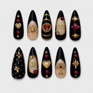 Può includere: Un set di dieci unghie finte a forma di mandorla con una palette di colori nero e nude. I design includono ciondoli dorati come cuori, stelle, soli e lune, accentuati da perline rosse e dorate, borchie metalliche e strisce dorate.