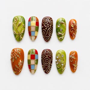 May include: A set of ten almond-shaped press-on nails featuring varied designs including green and orange marble patterns with gold leaf accents, a multi-coloured checkered grid, deep red swirls, and clear rhinestones set in gold metal filigree.