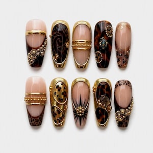 Könnte beinhalten: Ein Set aus zehn künstlichen Fingernägeln in Nude-, Braun- und Schwarztönen. Die Designs umfassen Leopardenmuster, goldene metallische Akzente, perlenartige Texturen und Sternmotive, die in zwei Reihen zu je fünf auf weißem Grund angeordnet sind.