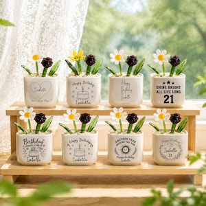 May include: Eight small, square, white ceramic pots with decorative floral arrangements. Each pot has a different message, including "Happy Birthday Isabella" and "Shine Bright All Life Long 21". The flowers include daisies and dark red roses.