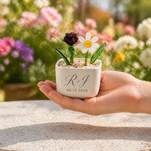 以下が含まれることがあります： ガラスの花のアレンジメントが入った、小さな正方形のオフホワイトのセラミックプランター。アレンジメントには、濃い赤色のバラと白いデイジーが含まれています。プランターには「R + J」と日付「06.15.2026」が刻まれています。