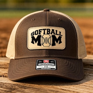 May include: Brown and tan trucker hat with the words "SOFTBALL MOM" in black block letters on a tan leather patch. The hat has a curved brim and a mesh back. The Richardson brand label is visible.