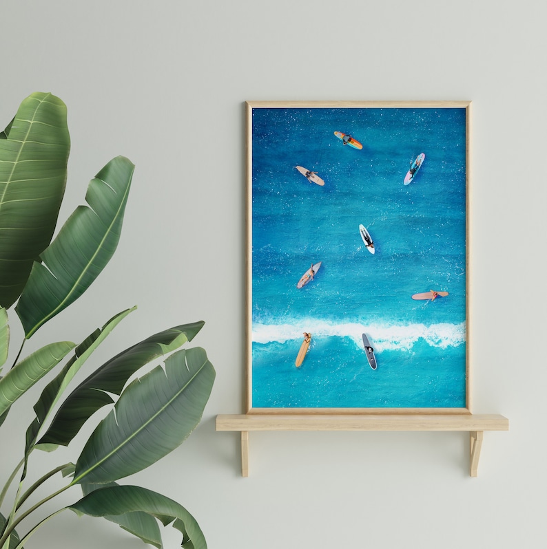 May include: A framed print of an aerial view of a turquoise ocean with multiple surfers on surfboards. The print is on a wooden shelf against a white wall.