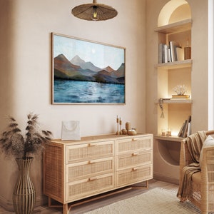 Puede incluir: Una escena interior serena con una pintura enmarcada de monta&ntilde;as y agua. Debajo de la obra de arte hay una c&oacute;moda de madera clara con cajones de rat&aacute;n tejido. Una l&aacute;mpara colgante tejida cuelga arriba y una silla tejida est&aacute; en la esquina. La habitaci&oacute;n tiene una paleta de colores neutros.