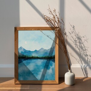May include: A framed watercolor painting of a mountain lake scene with a blue sky and a single white moon. The painting is in a wooden frame and is sitting on a white surface.