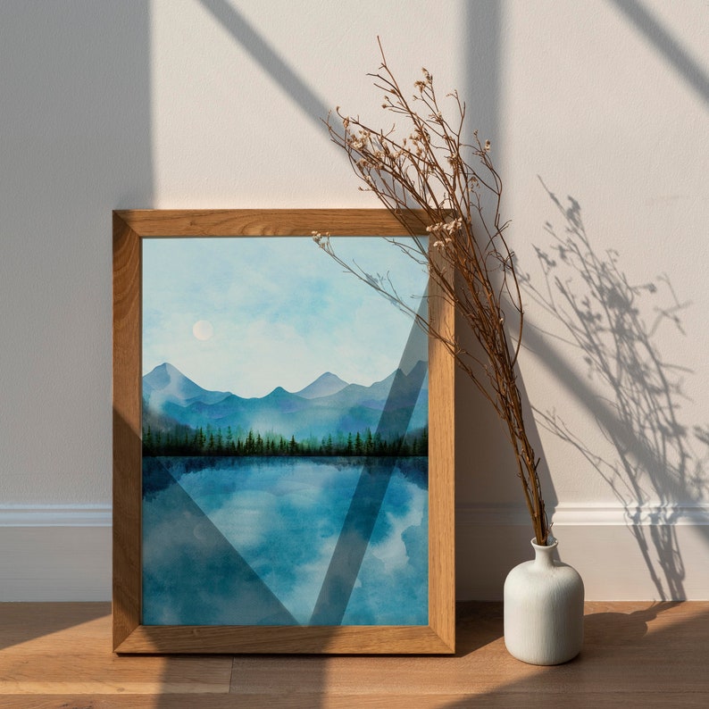 May include: A framed watercolor painting of a mountain lake scene with a blue sky and a single white moon. The painting is in a wooden frame and is sitting on a white surface.