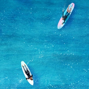 May include: Two people on surfboards in the ocean. The surfboards are white with pink and blue accents. The water is a bright blue.