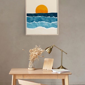 May include: A framed print of a minimalist watercolor painting depicting a sunset over the ocean. The painting features a large orange sun above a series of blue waves with white foam.