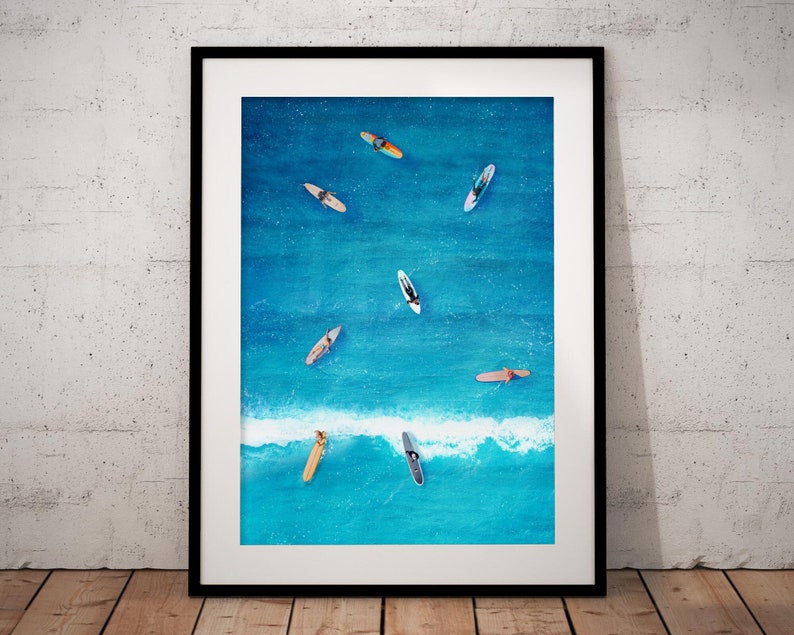 May include: An aerial view of a framed print depicting a turquoise blue ocean with seven surfers on surfboards. The surfers are all facing the same direction, and the waves are breaking in the background.