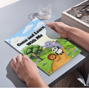 May include: A children's book titled "Come and Learn With Me" is held open, revealing colorful illustrations of an elephant, panda, and lion in a vibrant outdoor scene. The cover features a blue sky with clouds.