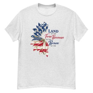 Peut inclure: Un t-shirt gris clair à manches courtes avec un motif de tournesol dont les pétales reprennent le drapeau américain. Le texte indique Land of the Free Because of the Brave en lettres bleues et rouges, mêlant écriture cursive et bâton.