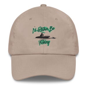 以下が含まれることがあります： ベージュ色の野球帽。つばがカーブしています。「I'd Rather Be Fishing」の文字が緑色の書体で、ボートから釣りをする人のグラフィックが描かれています。柔らかい素材で作られています。