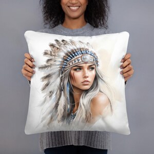 May include: Square white cushion featuring a watercolour portrait of a woman wearing a Native American headdress. The headdress is decorated with feathers, beads, and a central emblem. The woman has long, silver hair and a painted face.