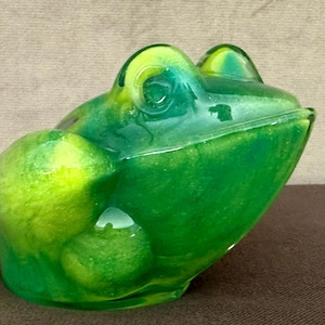 May include: A green and yellow frog-shaped trinket box. The box has a glossy finish and is made of a solid material. The frog's eyes and body are detailed, and the box is designed to open and close. The frog is sitting on a brown surface.