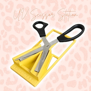 May include: A yellow UV scissor station with a pair of scissors. The scissors have black handles and silver blades. The text "UV Scissor Station" is written in a cursive font. The background is a light pink color with a leopard print pattern.