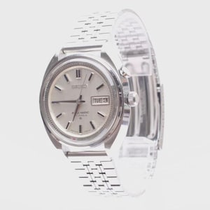 May include: A silver-toned Seiko wristwatch with a round face, metallic dial, and baton indices. The watch features a day-date window at the three o'clock position, a stainless steel link bracelet, and a crown on the right side of the case.