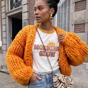 Könnte beinhalten: Eine grob gestrickte orangefarbene Strickjacke über einem weißen T-Shirt mit gelber Sonnengrafik und dem Text MORNING GLOW, kombiniert mit einer hellblauen Jeans und einer kleinen gemusterten Umhängetasche mit Kette sowie goldenen Ohrringen.