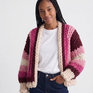 Può includere: Un cardigan a maglia grossa con righe orizzontali nei toni del crema, rosa chiaro, magenta e bordeaux scuro. Il capo presenta un design aperto sul davanti con un bordo spesso color crema lungo il colletto e i polsini, realizzato in filato spesso.