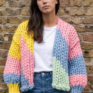 Op de afbeelding: Een grof gebreid vest met een kleurblokpatroon in geel, roze, lichtblauw en lichtgroen. Het kledingstuk is gemaakt van dik garen in een opvallende kabelsteek, met lange mouwen met geribbelde boorden en een open voorzijde.