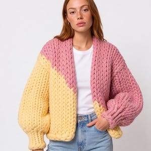 Könnte beinhalten: Eine grob gestrickte Strickjacke mit diagonalem Color-Block-Design in zartem Rosa und Hellgelb. Das Kleidungsstück hat eine offene Vorderseite, lange, voluminöse Ärmel mit gerippten Bündchen und eine dicke, strukturierte Garnkonstruktion.