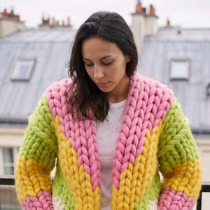 May include: A chunky knit cardigan featuring a color-blocked pattern of thick, oversized yarn in vibrant shades of pink, yellow, and lime green. The garment has a wide shawl collar and long sleeves, constructed with a prominent, heavy-gauge stitch.