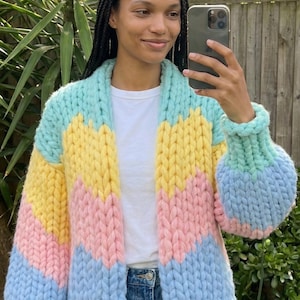 Op de afbeelding: Een chunky gebreid vest in pastelkleuren. De open trui heeft een chevron-ontwerp in lichtblauw, geel en roze. De mouwen zijn lang en hebben een afgeronde vorm. Het vest is gemaakt van dik garen.