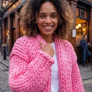 Könnte beinhalten: Eine leuchtend pinke Strickjacke aus grobem Strick mit dicker, übergroßer Garnstruktur, langen Ärmeln und offenem Vorderteil, getragen über einem schlichten weißen T-Shirt. Das Kleidungsstück besteht aus großen, sichtbaren ineinandergreifenden Garnschlingen.
