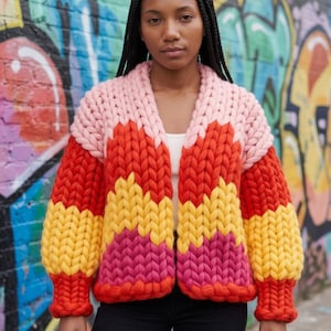 Puede incluir: Un cárdigan de punto grueso con un patrón de chevrón y bandas horizontales en rosa claro, rojo brillante, amarillo y magenta. La prenda tiene mangas largas con puños acanalados, un diseño frontal abierto y una textura de hilo grueso y voluminoso.