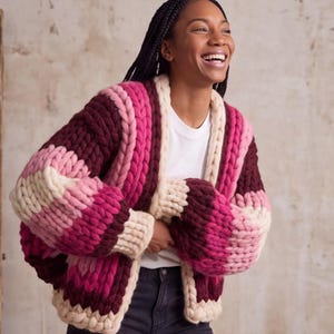 May include: A chunky knit cardigan featuring thick, horizontal stripes in shades of cream, light pink, magenta, and deep burgundy. The garment has a relaxed, open-front design with long sleeves and a thick, textured yarn construction throughout.