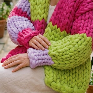 Könnte beinhalten: Ein grob gestrickter Cardigan in leuchtenden Farben. Der übergroße Pullover hat große Maschen und ein Colorblock-Design mit Pink-, Grün- und Lilatönen. Das gemütliche, handgefertigte Kleidungsstück ist perfekt, um jedem Outfit einen Farbtupfer zu verleihen.