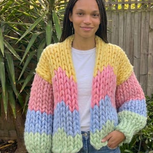 Pode incluir: Um cardigã de malha grossa com listras horizontais coloridas em amarelo, rosa, azul claro e verde claro. A peça possui mangas compridas, frente aberta e uma construção de fio espesso e texturizado com um padrão de pontos visível.