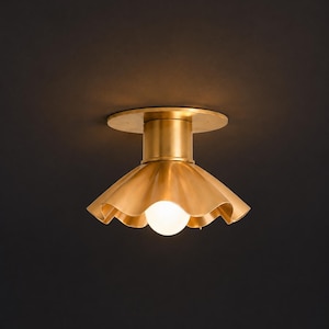 Può includere: Un lampada da soffitto dorata con un paralume a balze e una lampadina bianca rotonda. L'apparecchio ha una base cilindrica e una piastra di montaggio circolare. Lo sfondo è scuro.