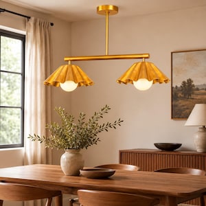 May include: A gold-toned metal linear chandelier featuring two downward-facing, pleated, bell-shaped shades. Each shade houses a single, spherical, frosted white light bulb. The fixture is suspended from the ceiling by a central vertical rod.