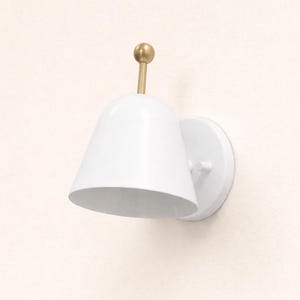 May include: White wall sconce with a conical shade and a brass-colored accent. The sconce has a round base and a small, cylindrical arm. The design is minimalist and modern, suitable for various interior styles.