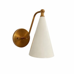 May include: A wall-mounted sconce with a conical white shade and a brass-colored arm and mounting plate. The fixture has a minimalist design, suitable for various interior styles. The shade is smooth and opaque.