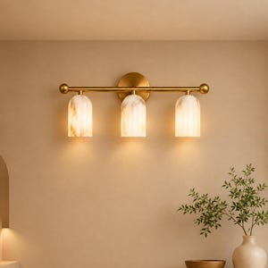 May include: A wall-mounted light fixture featuring a horizontal brass-toned metal bar with three downward-facing, arched, translucent white and beige marbled glass shades. The fixture is attached to the wall with a circular brass-toned backplate.