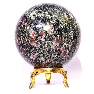 Puede incluir: Una esfera decorativa con un patrón moteado negro, verde y rojo, apoyada sobre un soporte dorado de tres patas. La esfera tiene una superficie pulida y reflectante, y el soporte tiene un diseño detallado y ornamentado. La estética general es elegante y artística.