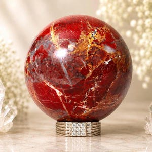 Puede incluir: Una piedra de jaspe rojo pulida y esférica con intrincados patrones de vetas doradas, blancas y gris oscuro. La esfera se apoya sobre un anillo de metal plateado que muestra un engaste cuadrado con nueve piedras pequeñas y claras.