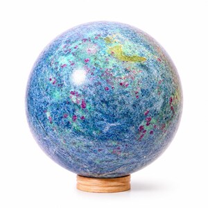 May include: A polished spherical stone orb featuring a mottled texture of deep blue, light blue, and green mineral inclusions, accented with scattered vibrant magenta-pink spots, resting on a small, circular, light-colored wooden base.