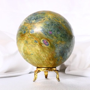 May include: A polished spherical stone featuring a mottled surface with shades of olive green, golden yellow, and deep blue, accented by small, vibrant magenta inclusions. The sphere rests on a small, three-legged metallic gold-toned stand.
