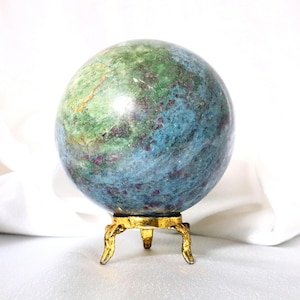 May include: A polished spherical stone featuring a mottled surface with vibrant shades of green, blue, and deep red speckles. The sphere rests securely upon a small, ornate, three-legged gold-colored metal stand with a textured finish.