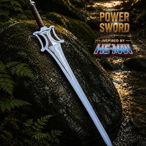 Può includere: Una spada in metallo argentato con un'impugnatura avvolta in marrone e una guardia dalla forma distintiva. La lama è lunga e appuntita. Il testo sull'immagine recita: POWER SWORD INSPIRED BY HE-MAN. La spada è appoggiata contro una grande roccia muschiosa.
