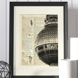 May include: Black framed print featuring a vintage dictionary page with a grayscale image of the Berlin TV Tower. The print has a white border and is displayed on a wooden surface with decorative items.